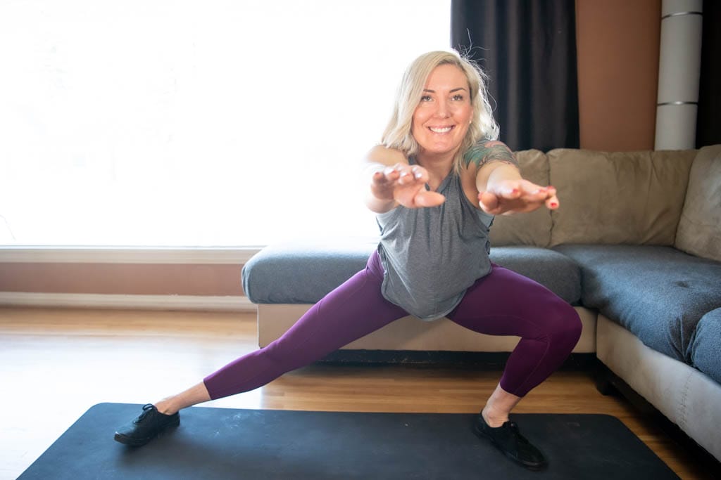 A blond woman performs a cossack squat in her living room as part of an at-home fitness program.