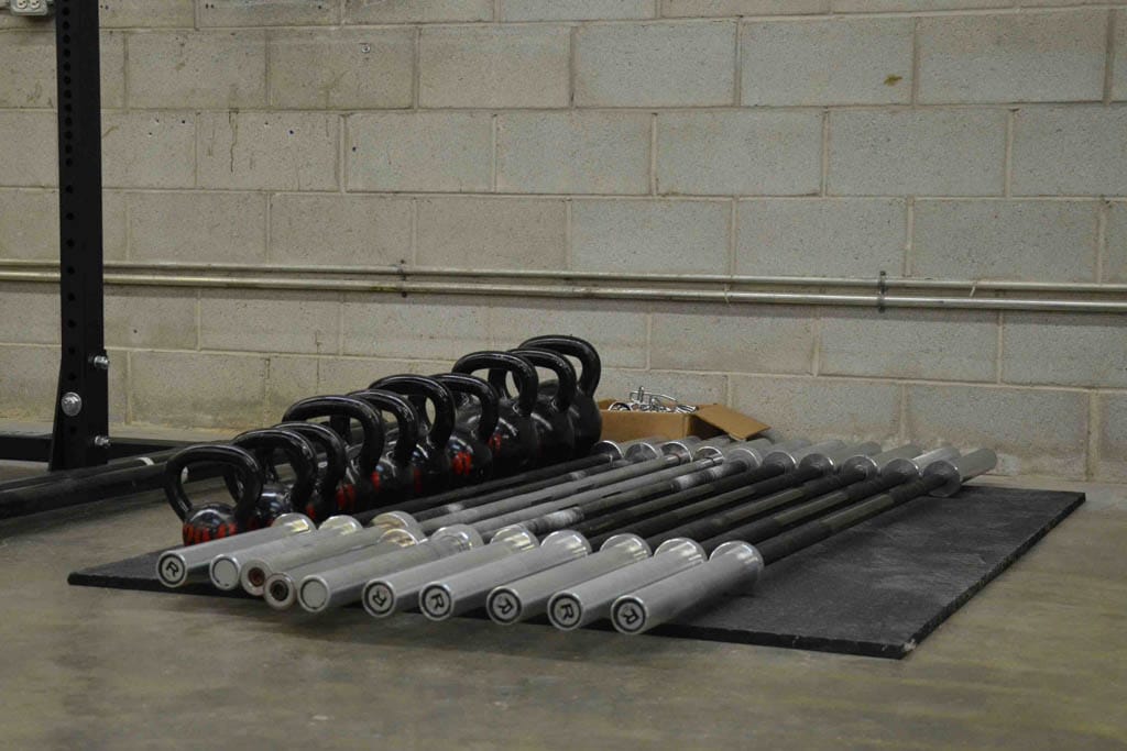 A pile of kettlebells and barbells lie on a piece of black rubber in a warehouse gym.