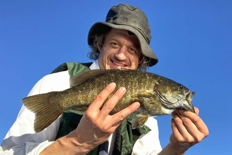 A smiling man holds a large bass caught on Black Sturgeon after an online fitness workout.