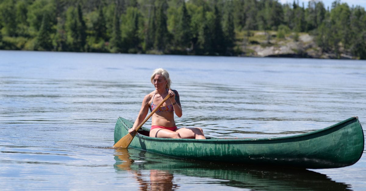 A blond woman in a bikini paddles a canoe as part of an online fitness workout.