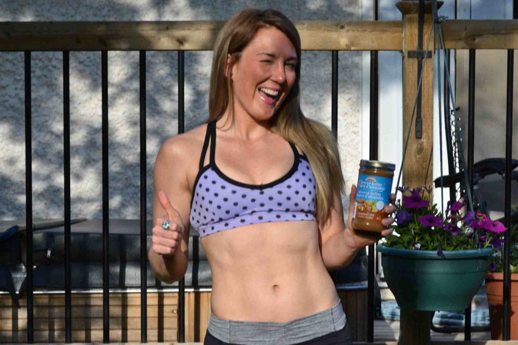 A fit woman in a tank top holds a jar of peanut butter and gives the thumbs up sign.