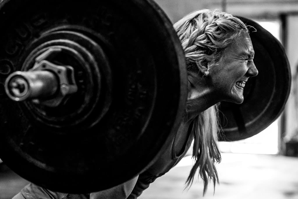 A closeup image of a woman gritting her teeth while squatting with heavy barbell on her back.